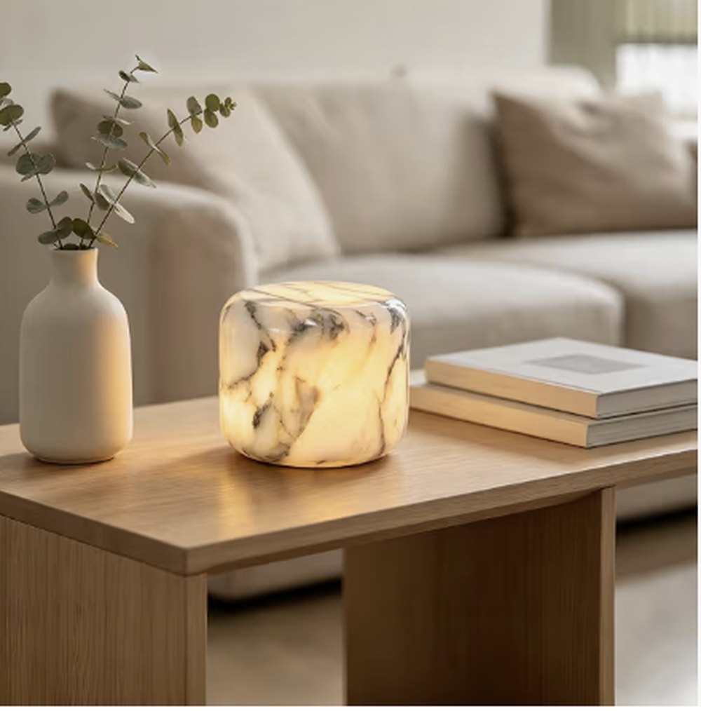 A small marble table lamp glowing warm on a wooden coffee table beside a vase of eucalyptus and two books.