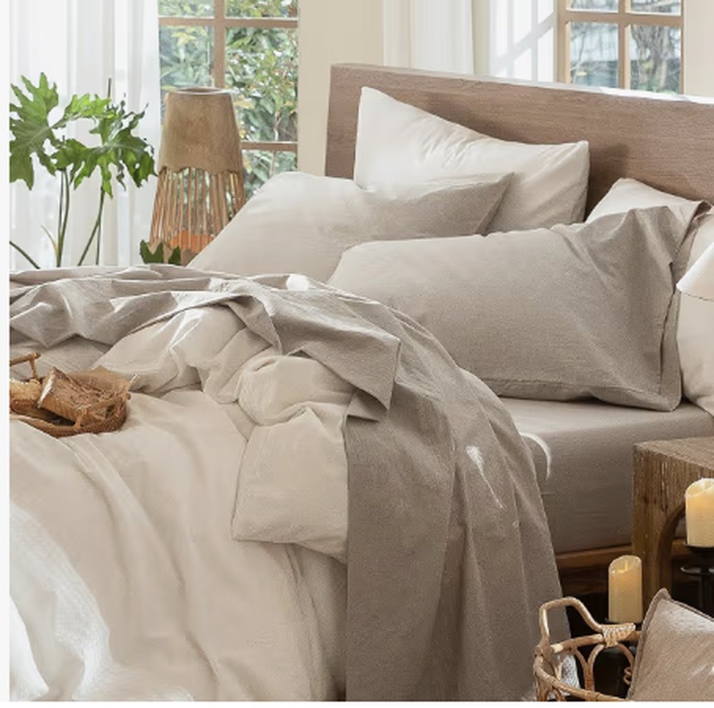 A bed made up in washed cotton sheets in oatmeal and stone, with a wooden headboard and a candle beside it.
